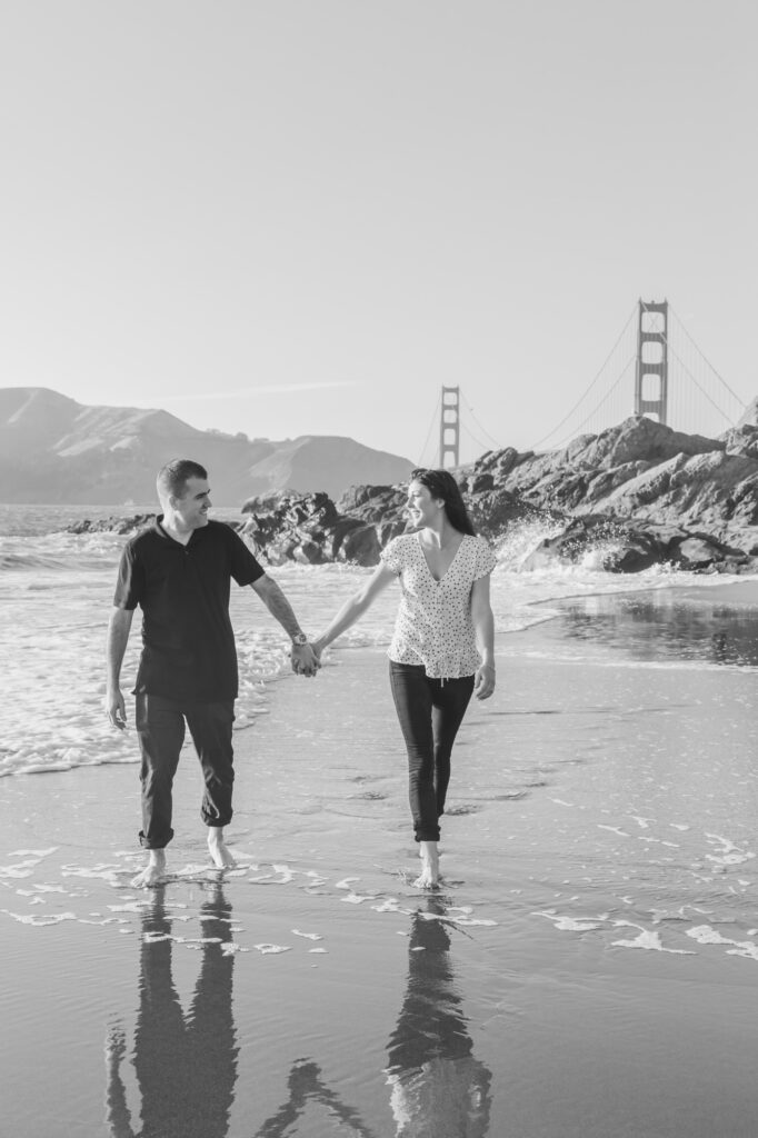 couple holding hands walking along the shoreline at Baker Beach with the Golden Gate Bridge in the background during a San Francisco engagement session