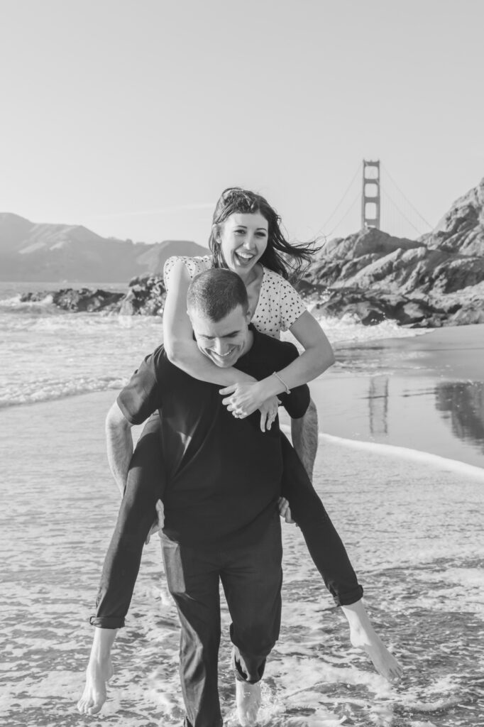 playful couple at Baker Beach with piggyback moment near the shoreline and Golden Gate Bridge in the background during a San Francisco engagement session