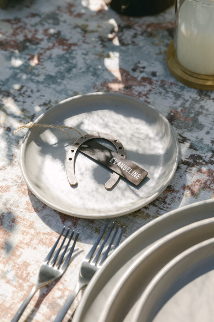 Elegant western wedding place setting featuring layered plates, linen napkin, and horseshoe place card on patterned tablecloth at Holman Ranch.