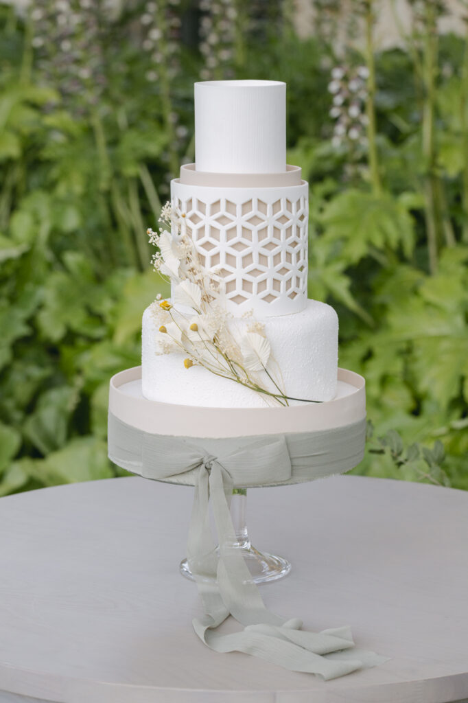 Modern white tiered western wedding cake with textured finish, geometric lattice detail, and delicate dried florals displayed on pedestal at Holman Ranch in Carmel Valley, California.