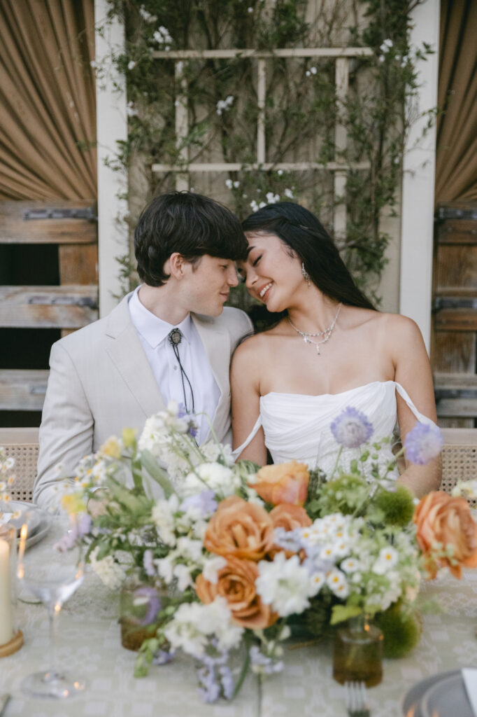 Bride and groom seated closely at reception table surrounded by lush floral arrangements in soft orange, lavender, and white tones at luxury western wedding at Holman Ranch in Carmel Valley.