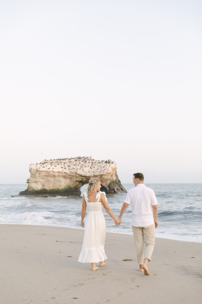 couple walking along the shoreline at Natural Bridges State Beach with the iconic rock formation in Santa Cruz California