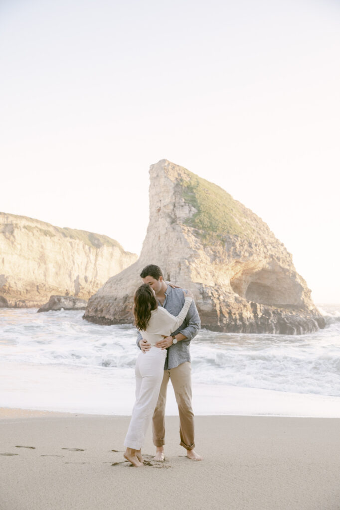 couple embracing on beach with shark fin rock and cave at Shark Fin Cove engagement session in California