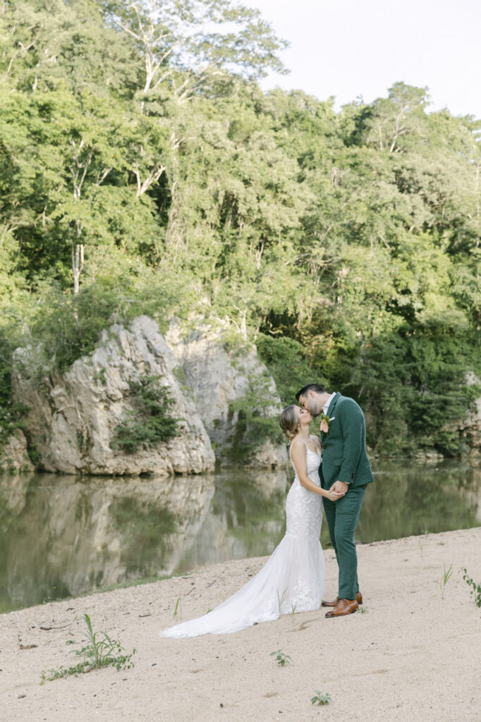 Bride and groom kissing on a sandy riverbank framed by limestone rock and rainforest, luxury Belize destination wedding photographer Paulina Perrucci