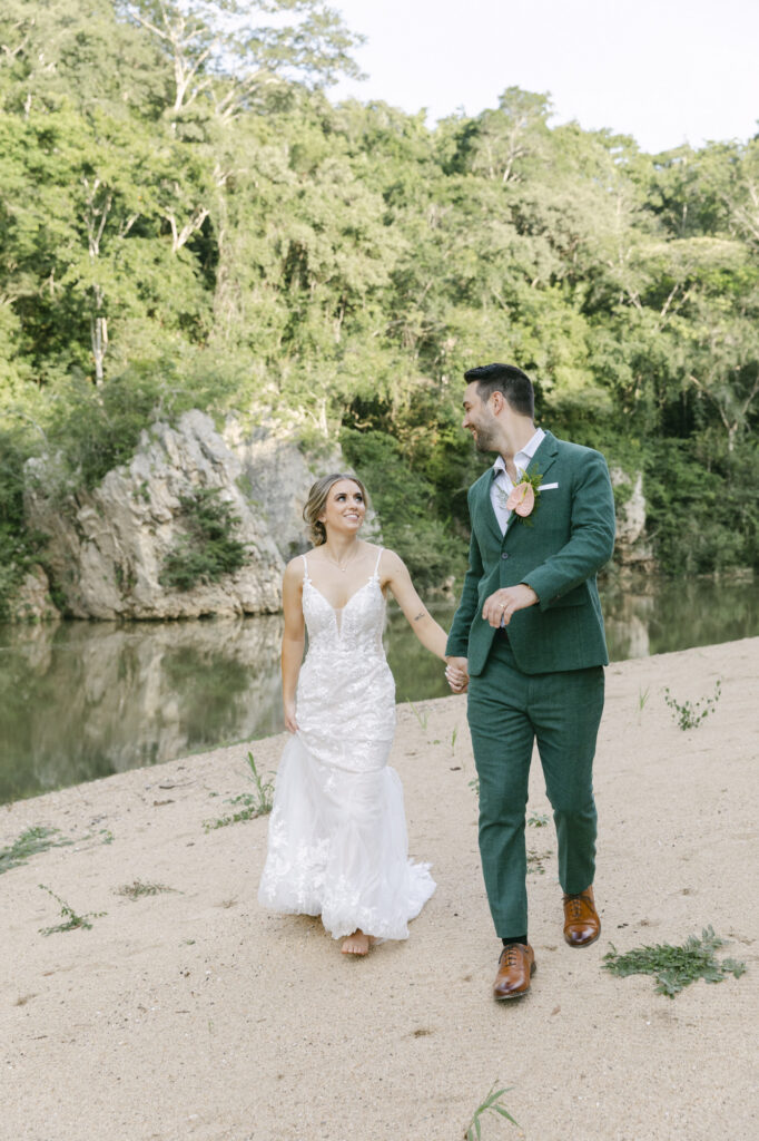 Bride and groom walking hand in hand along a sandy riverbank framed by limestone and rainforest, luxury Belize destination wedding photographer Paulina Perrucci
