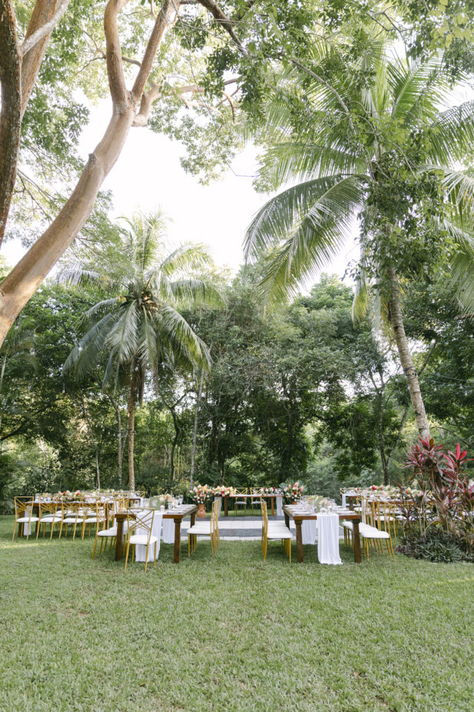 Outdoor jungle wedding reception with long wood tables, gold chairs and tropical florals beneath the palms, Belize destination wedding photographer Paulina Perrucci