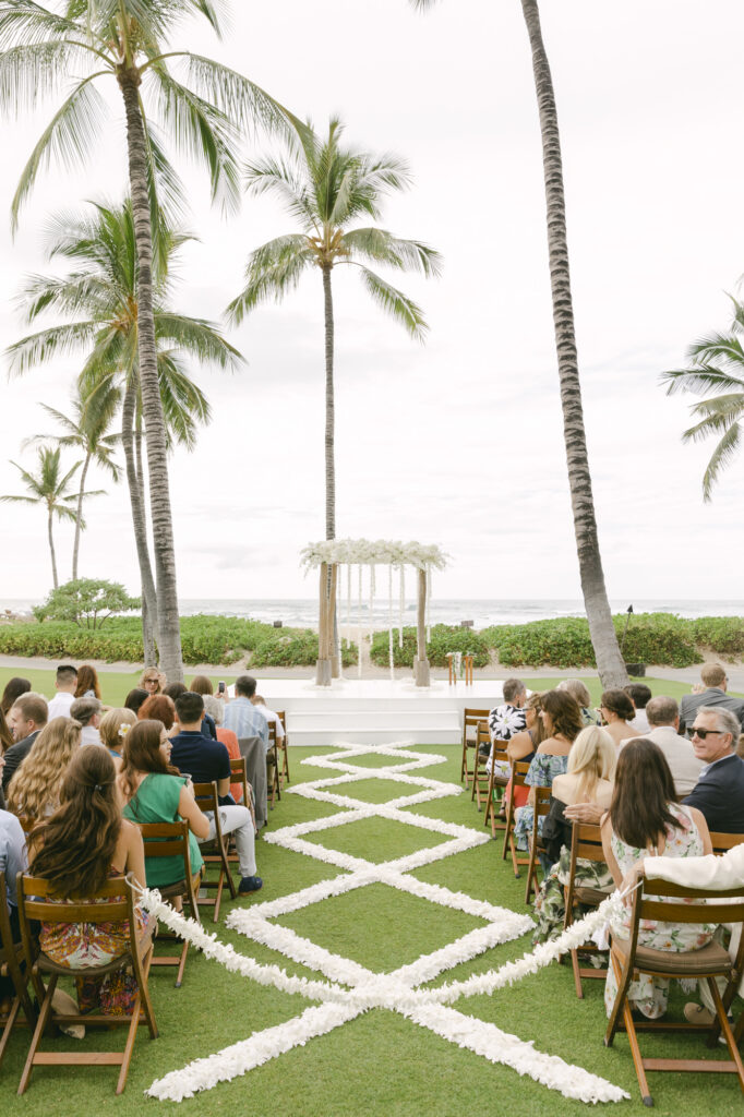 Outdoor wedding ceremony at Four Seasons Hualalai with guests seated on a lawn, geometric white petal aisle design, tall floral arch altar, palm trees, and ocean views