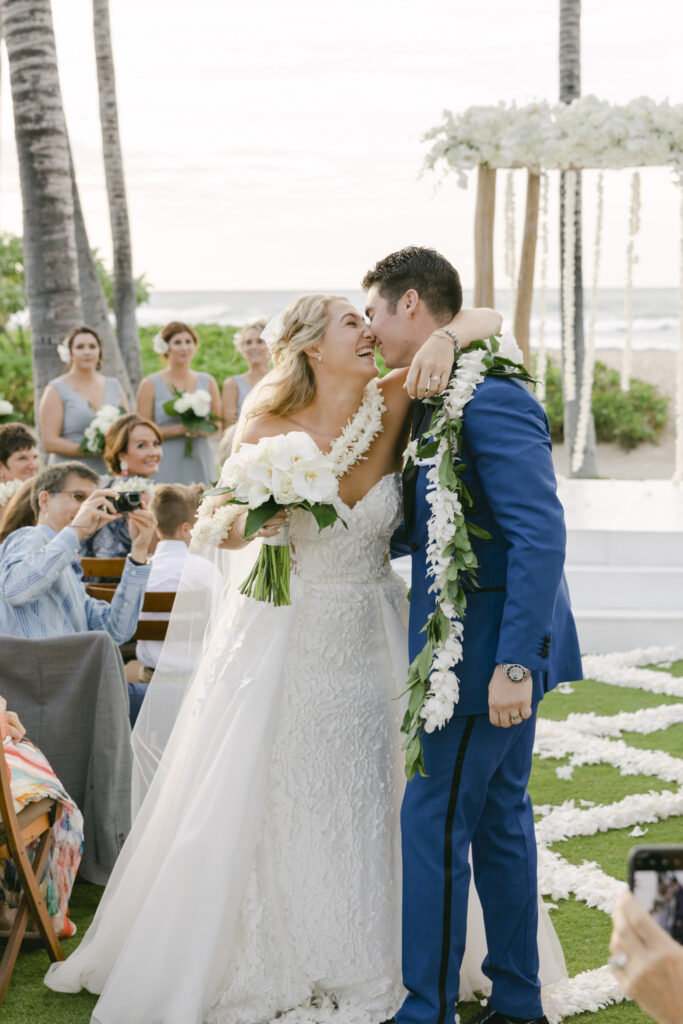 Bride and groom laughing together after their ceremony at Four Seasons Hualalai, bride in a lace sweetheart ballgown and groom in a cobalt blue suit wearing a traditional Hawaiian orchid lei