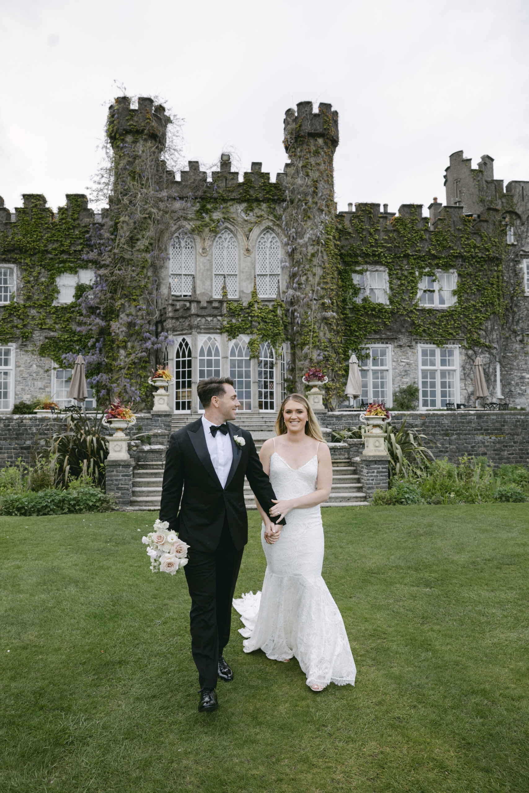 Bride and groom walk hand in hand on the lawn at Luttrellstown Castle, with wisteria climbing the stone walls behind them.