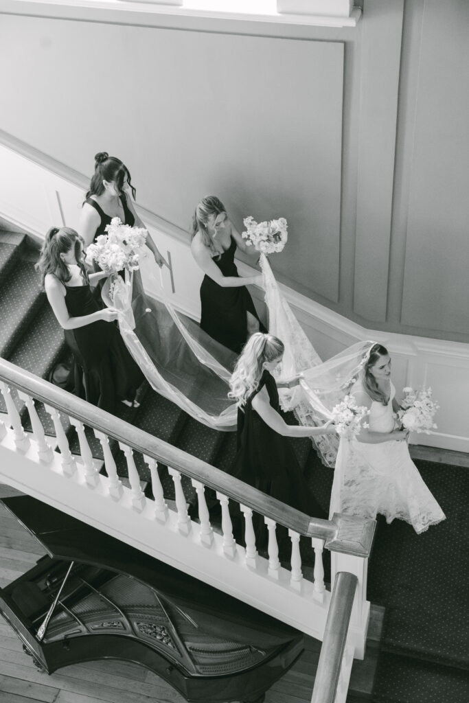 Black and white image of the bride descending the grand staircase at Luttrellstown Castle as bridesmaids carry her veil.