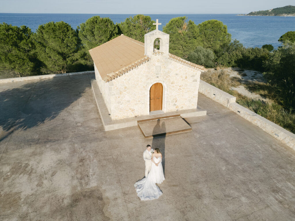 Drone view of a bride and groom outside a stone chapel overlooking the ocean during a luxury Zakynthos Greece destination wedding with editorial style wedding photography.
