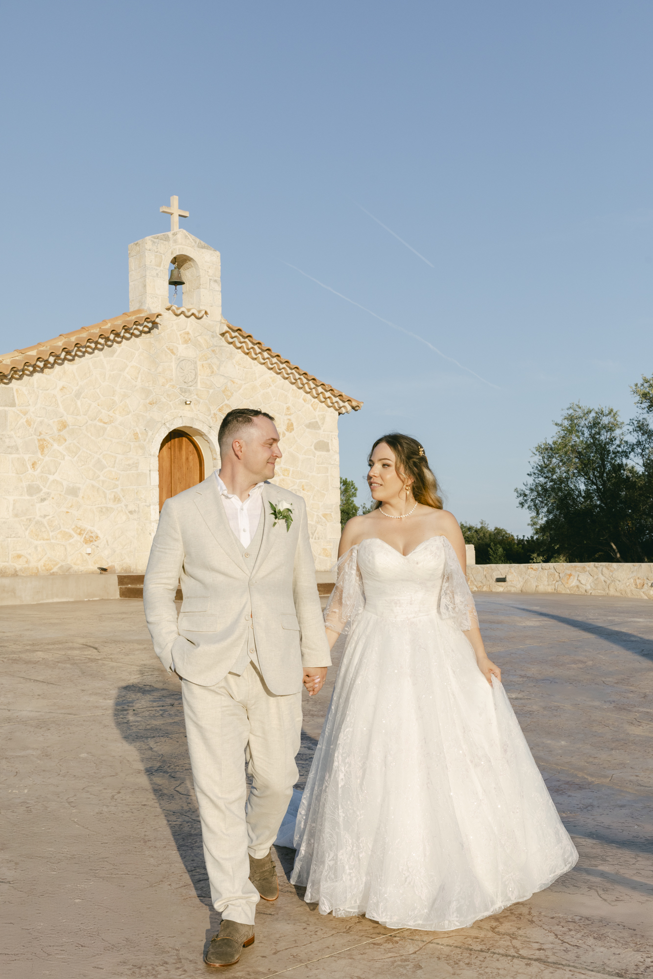 Bride and groom walking hand in hand in front of a stone chapel during a luxury Zakynthos Greece destination wedding with editorial style wedding photography.