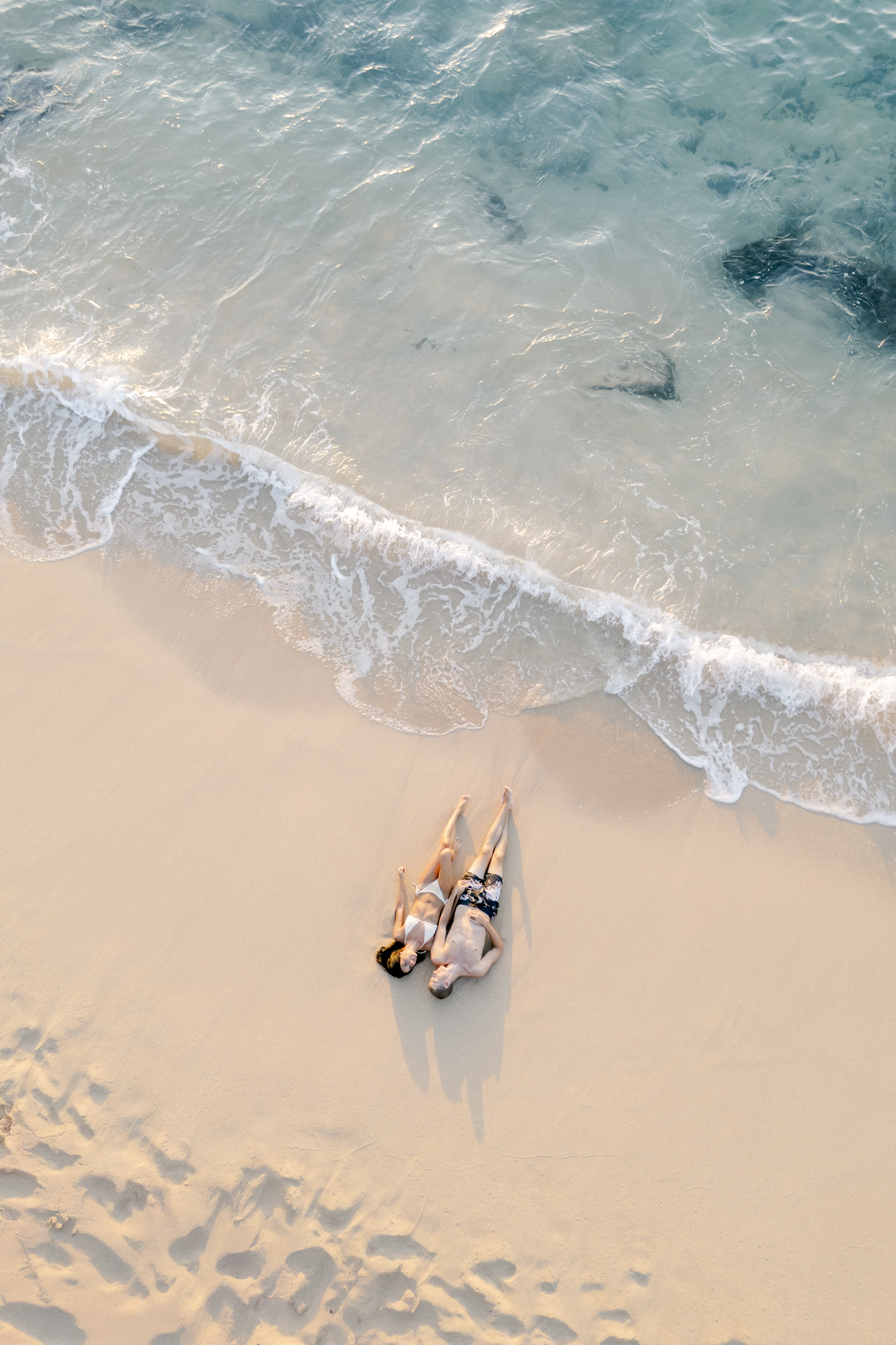 Aerial view of a couple lying on a sandy Hawaiian beach at the water's edge during their engagement session