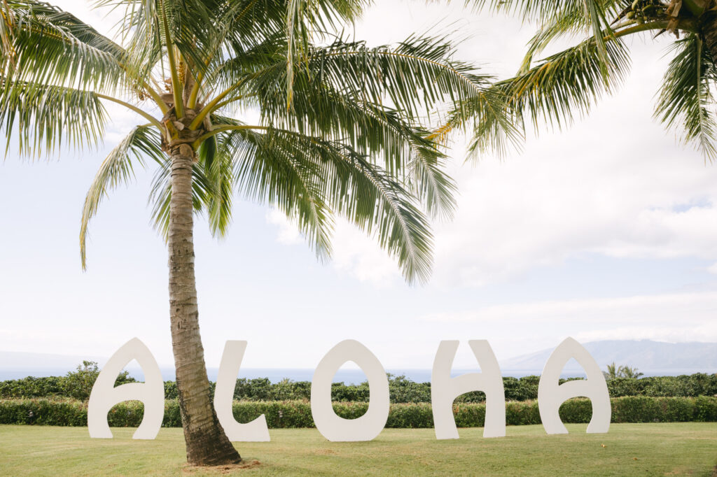 Large white Aloha letter sign on the lawn of a private estate wedding in Maui Hawaii with a palm tree and sweeping ocean and mountain views in the background