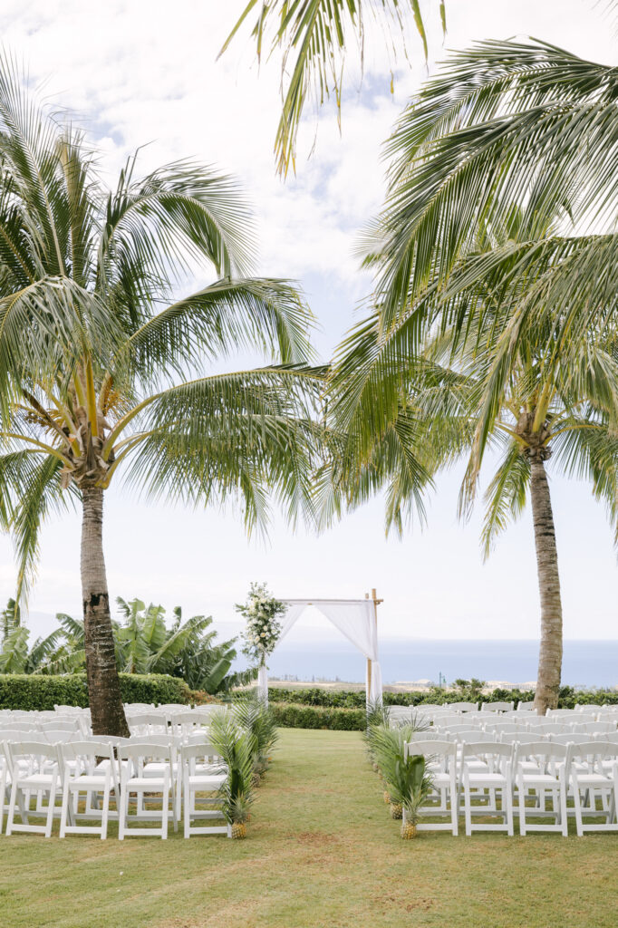 Outdoor ceremony setup at a Maui private estate wedding with wooden bamboo arch, white florals, and fresh pineapples lining the aisle with ocean views in the background