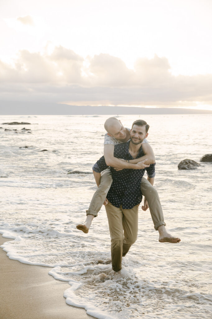 Gay couple laughing during a piggyback ride through the ocean waves on the beach during their Maui engagement session at sunset