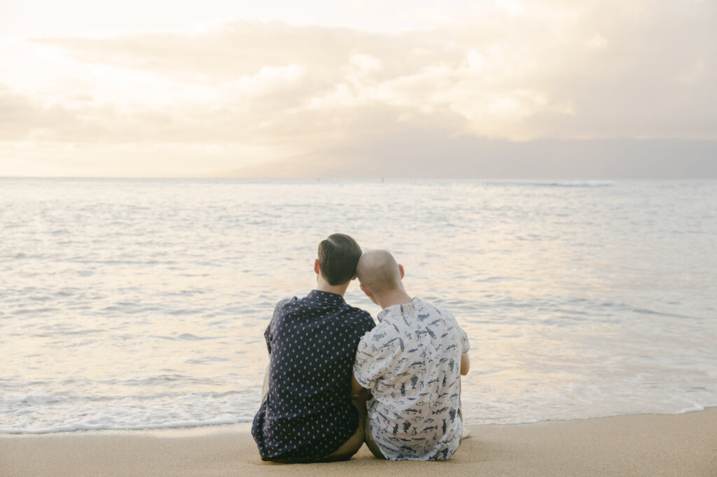 Gay couple sitting together on a Maui beach watching the sunset during their engagement session