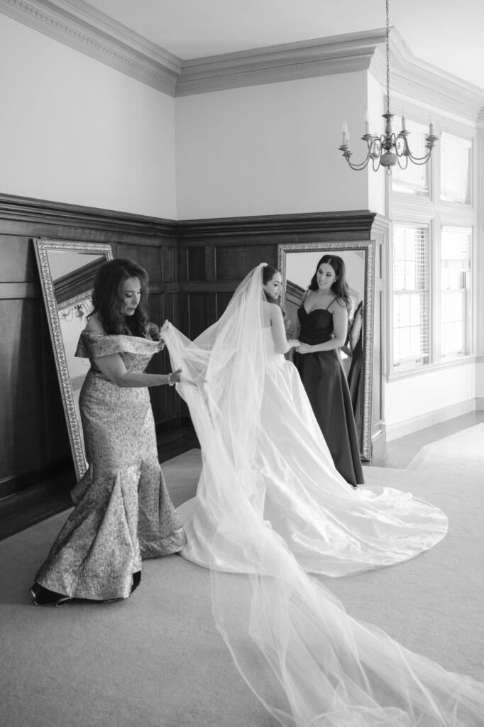 Black and white photo of the mother of the bride arranging the cathedral veil in a getting-ready room at Kohl Mansion with a large mirror reflecting the moment