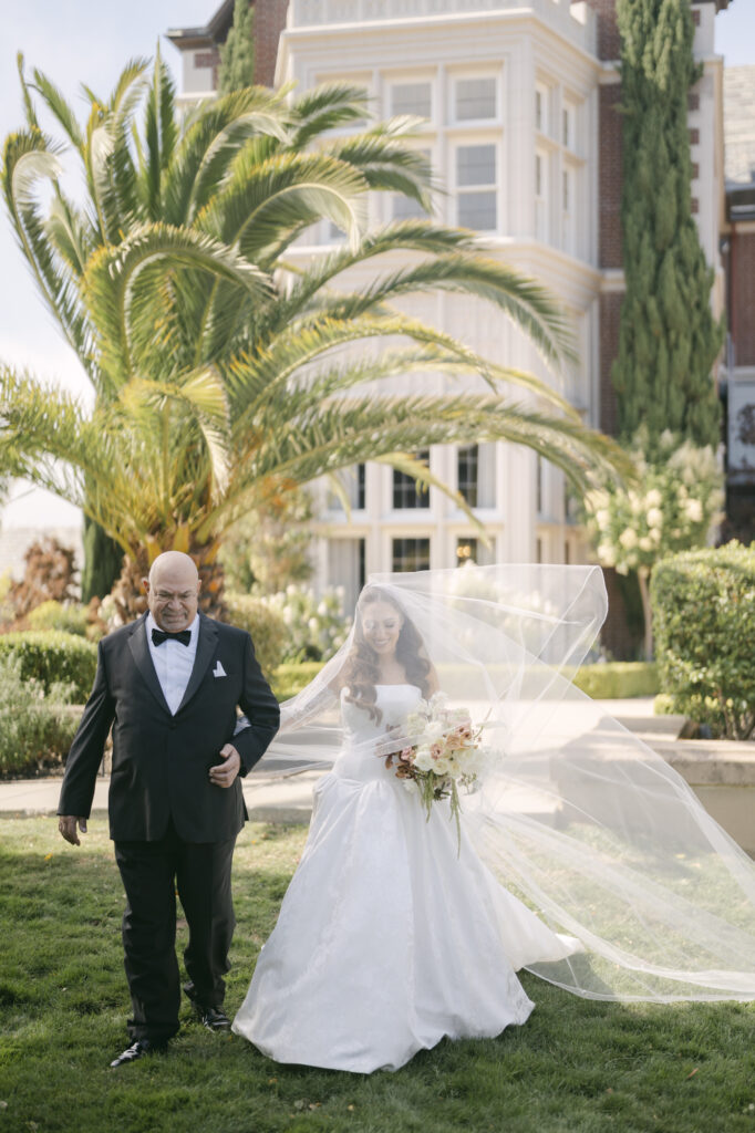 Father walking the bride down the outdoor lawn aisle at Kohl Mansion with her cathedral veil billowing in the breeze and the brick mansion facade and palm trees in the background