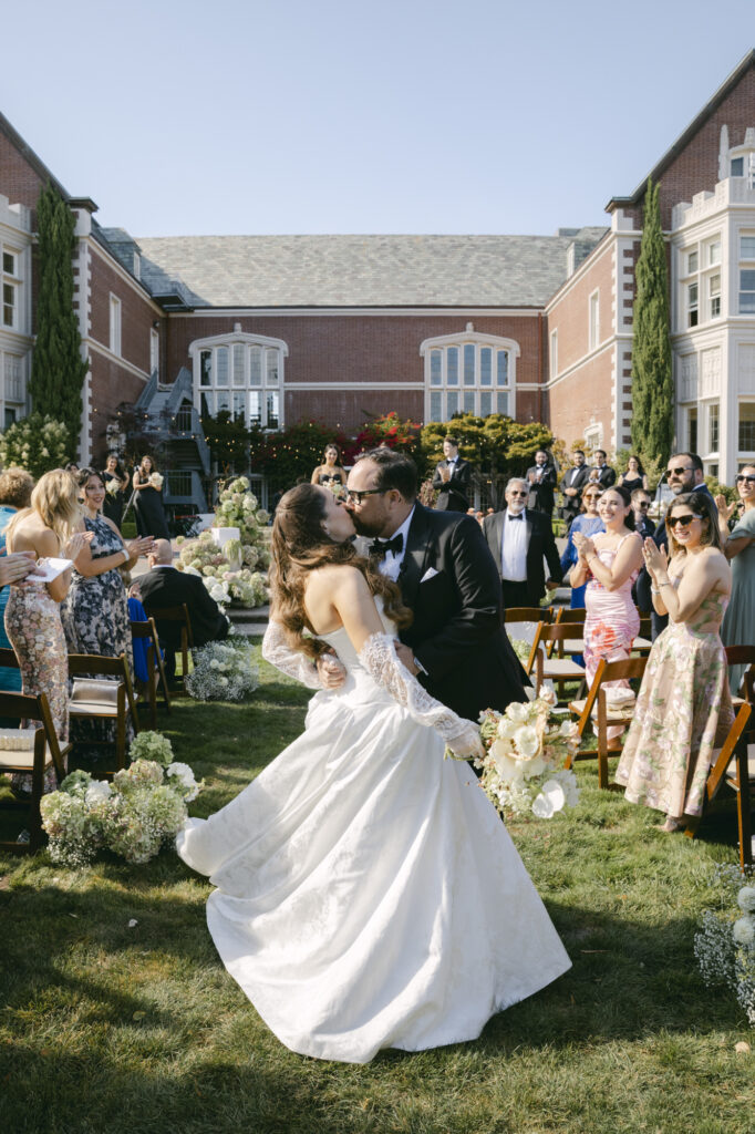 Bride and groom sharing their first kiss during the outdoor lawn ceremony at Kohl Mansion as guests cheer from wooden chairs on the grounds