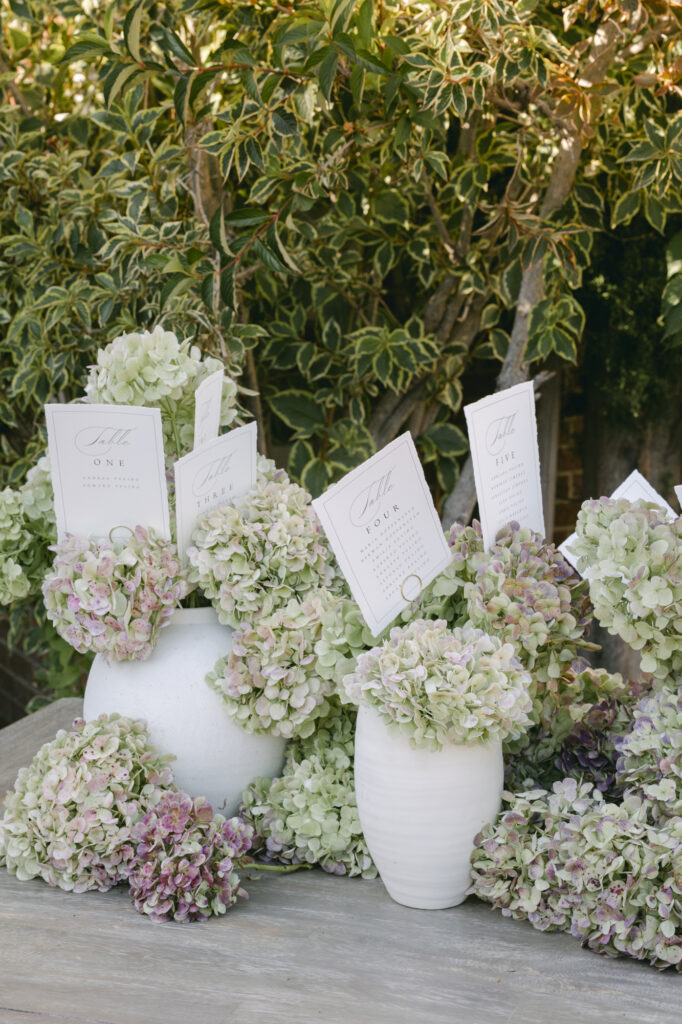 Escort card seating chart display at a Kohl Mansion wedding featuring blush and green hydrangeas overflowing from white ceramic urns with calligraphed table cards for a luxurious wedding.