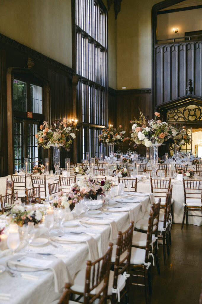 Wide view of the Kohl Mansion Great Hall reception with long farm tables, ivory linens, chiavari chairs, and tall crystal vase centerpieces with peach and blush florals for a luxurious wedding. 