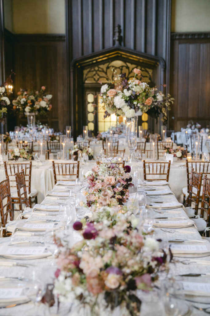 Candlelit farm table at a Kohl Mansion reception with peach, blush, and burgundy low floral centerpieces and dark oak paneled walls in the background for a luxurious wedding. 