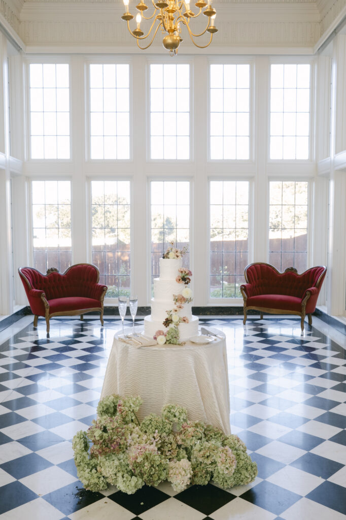 Four-tier white wedding cake surrounded by green hydrangeas on the black and white checkered marble floor of the Kohl Mansion conservatory with red velvet chairs in the background