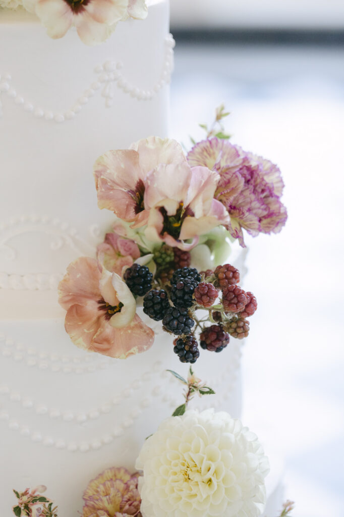 Close-up of blush poppies, mauve carnations, and fresh blackberries cascading down the tiers of the white wedding cake at a Kohl Mansion wedding