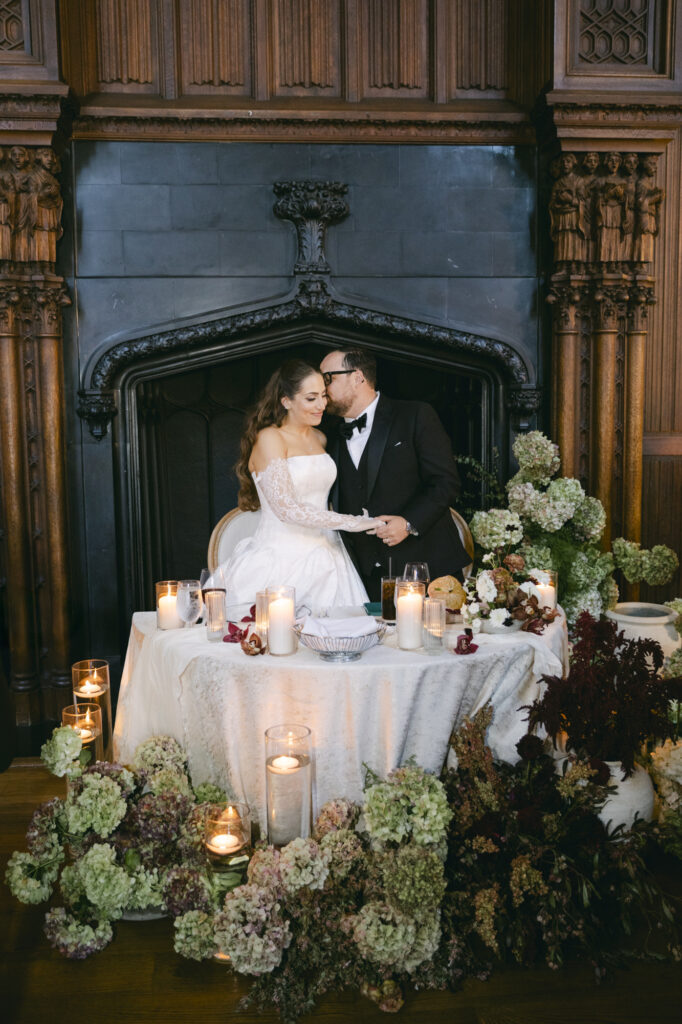 Bride and groom at their sweetheart table in front of the carved Gothic stone fireplace at Kohl Mansion surrounded by a lush floral installation and pillar candles