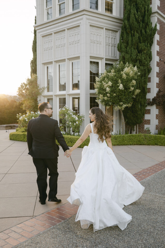 Bride and groom walking hand in hand at golden hour with the illuminated white Gothic tower of Kohl Mansion glowing behind them at sunset