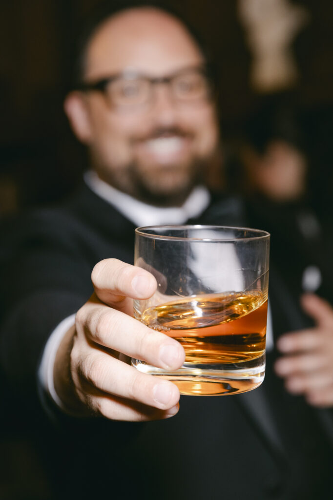 Groom smiling and holding a rocks glass of whiskey at the speakeasy bar during a Kohl Mansion wedding reception