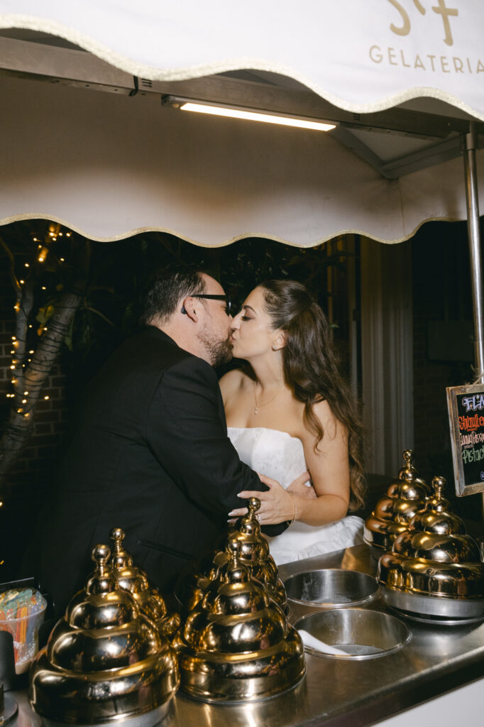 Bride and groom sharing a kiss at the gelateria cart under string lights during their Kohl Mansion wedding reception with the Gelateria sign in the background