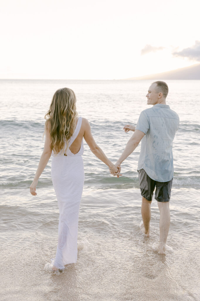 Couple walking hand in hand into the ocean at sunset during a Maui Hawaii engagement session with soft golden light and a mountain in the background