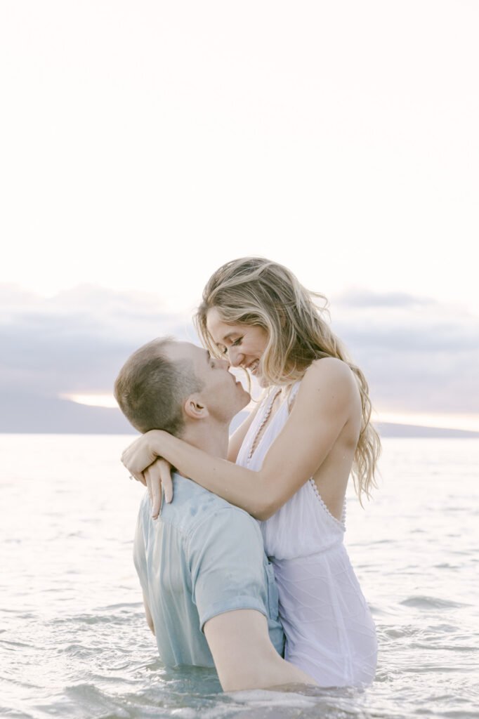 Couple laughing and embracing while standing in the ocean during a golden hour Maui Hawaii engagement session with a soft pastel sky