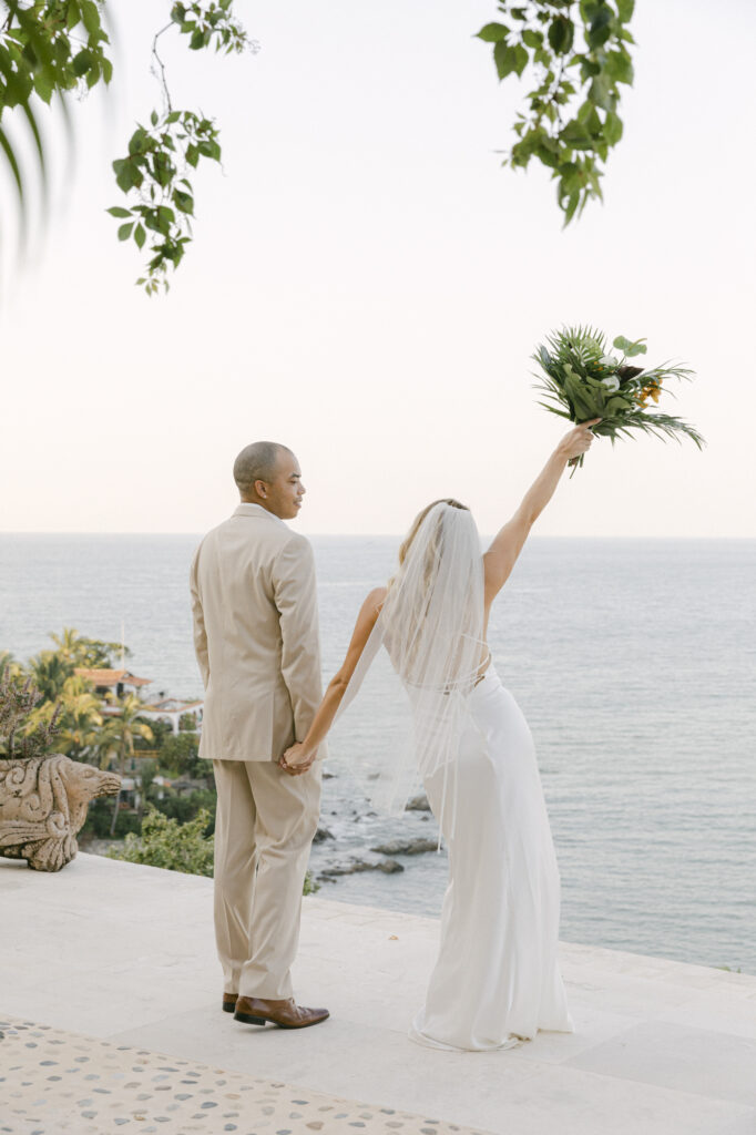 Bride and groom from behind on a rooftop terrace at La Gran Villa Sayulita, bride raising her tropical bouquet in celebration with the Sayulita coastline and Pacific Ocean below