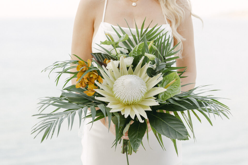 Close-up of a tropical bridal bouquet featuring a white king protea, golden yellow cymbidium orchids, anthurium leaves, and palm fronds held by a bride in a silk slip gown at a Sayulita Mexico wedding