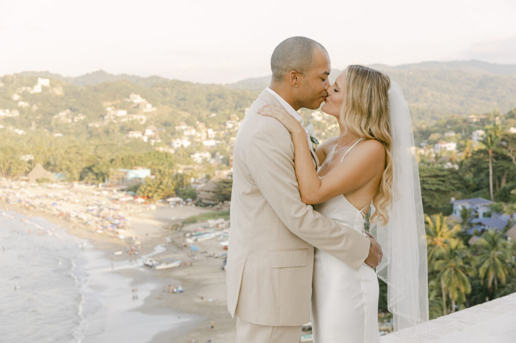 Bride and groom kissing on a rooftop overlooking the beach town and coastline of Sayulita, Mexico at golden hour, bride in a silk slip gown with veil and groom in a tan suit