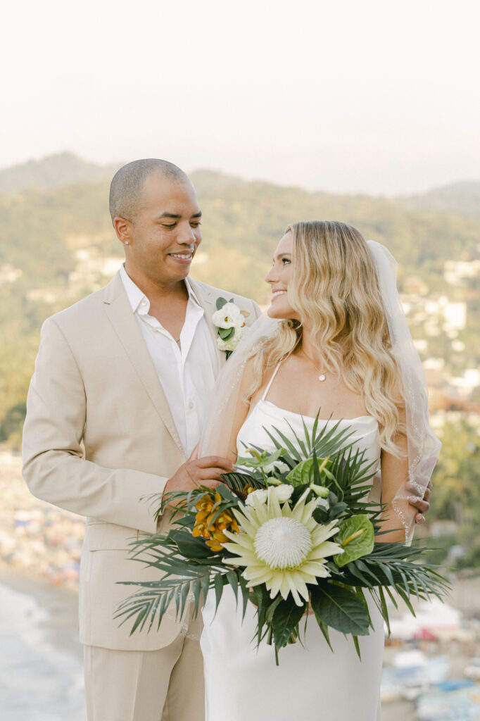 Bride and groom portrait at La Gran Villa Sayulita overlooking the colorful hillside town and coastline, bride holding a tropical king protea and palm frond bouquet in a silk slip gown