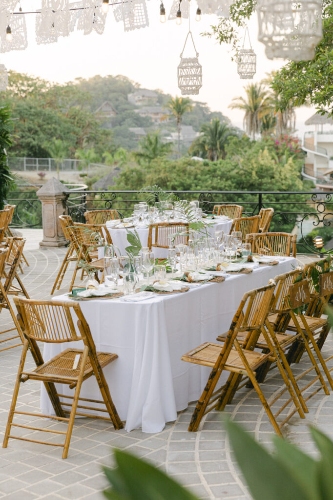 Outdoor wedding reception long table with white linens, bamboo rattan chairs, greenery centerpieces, papel picado, and hanging lanterns at La Gran Villa Sayulita with jungle and hillside views