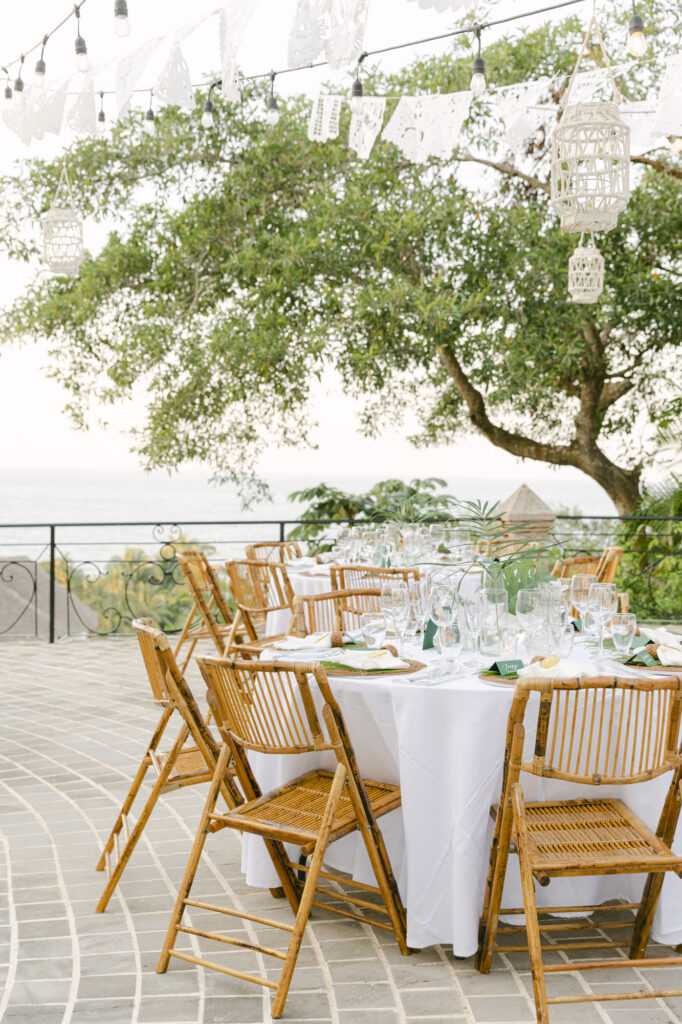 Outdoor wedding reception tablescape at a Sayulita Mexico wedding with bamboo rattan chairs, white linens, papel picado banners, hanging birdcage lanterns, and ocean views