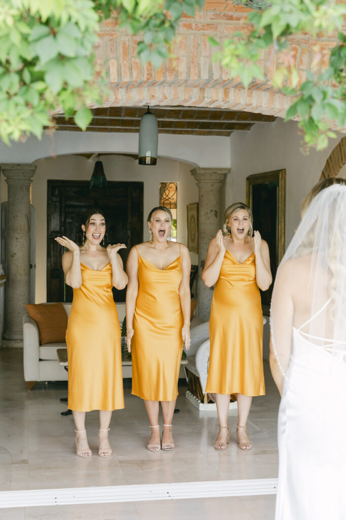 Three bridesmaids in golden amber satin dresses reacting with joy and surprise to seeing the bride for the first time at a La Gran Villa Sayulita wedding