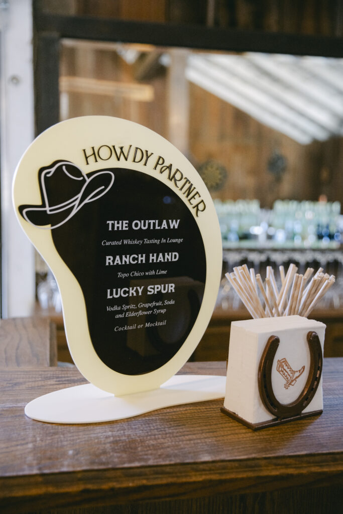 Western-themed acrylic cocktail menu sign reading Howdy Partner with drink options including The Outlaw, Ranch Hand, and Lucky Spur, displayed on a wooden bar at Stonepine Estate