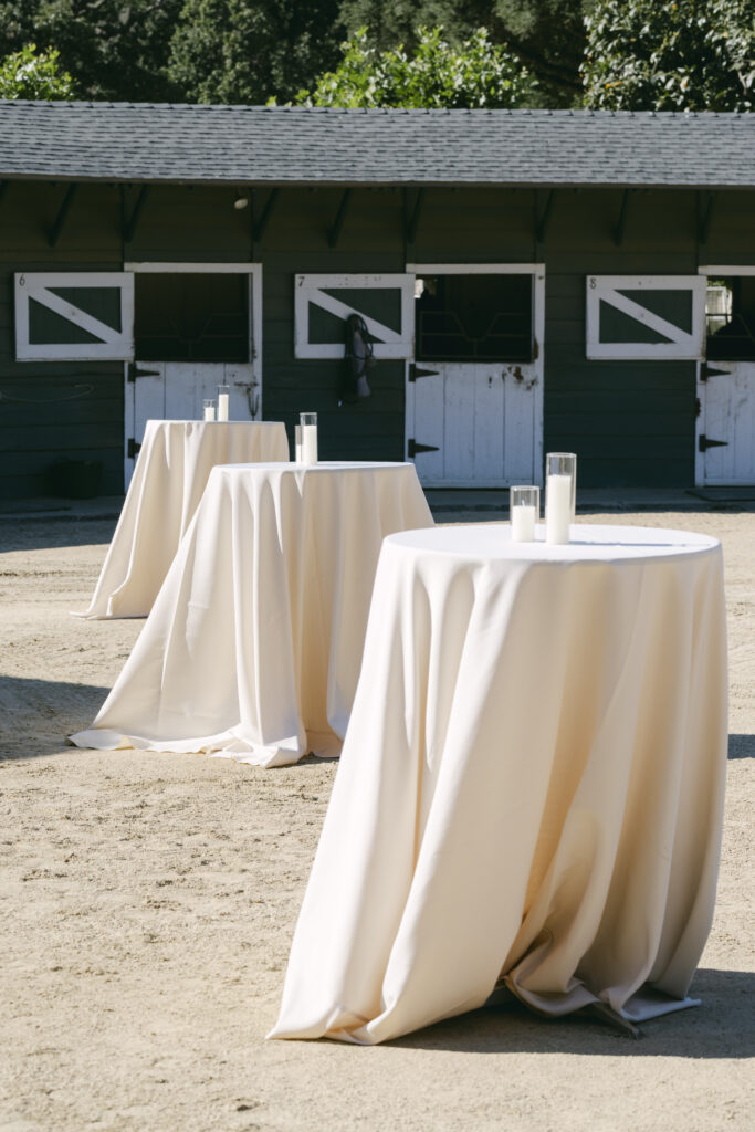 Cream-draped cocktail tables with candles set up in the sandy courtyard in front of the green horse stables at Double H Ranch at Stonepine Estate in Carmel Valley