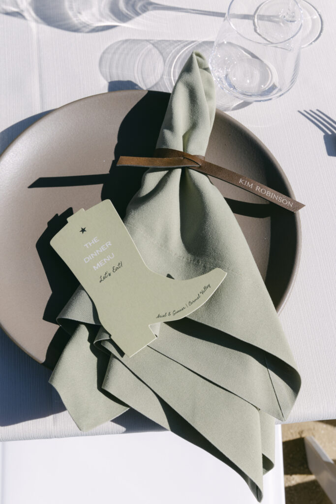 Close-up of a rehearsal dinner place setting at Stonepine Estate with a sage green folded napkin, brown ribbon, personalized place card reading Kim Robinson, and a printed menu card