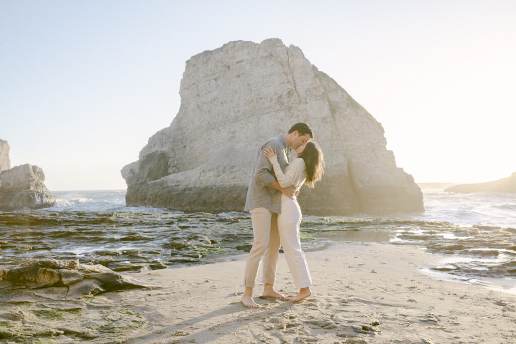 Couple sharing a passionate kiss on the beach at Shark Fin Cove with the dramatic shark fin rock formation at golden hour in Davenport