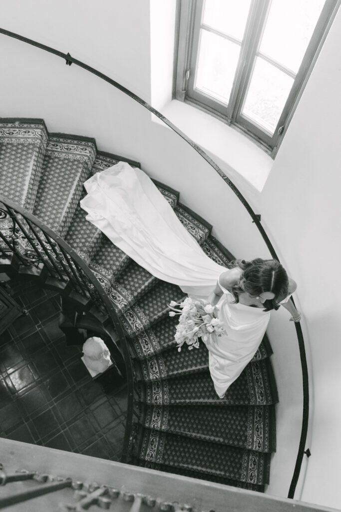 Black and white aerial view looking down at a bride descending the ornate spiral staircase inside Chateau Noel at Stonepine Estate, her wedding gown and veil fanning out across the patterned carpet steps