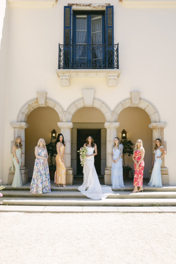 Bride and bridesmaids in colorful and pastel gowns standing on the stone steps beneath the arched entryway of Chateau Noel at Stonepine Estate in Carmel Valley