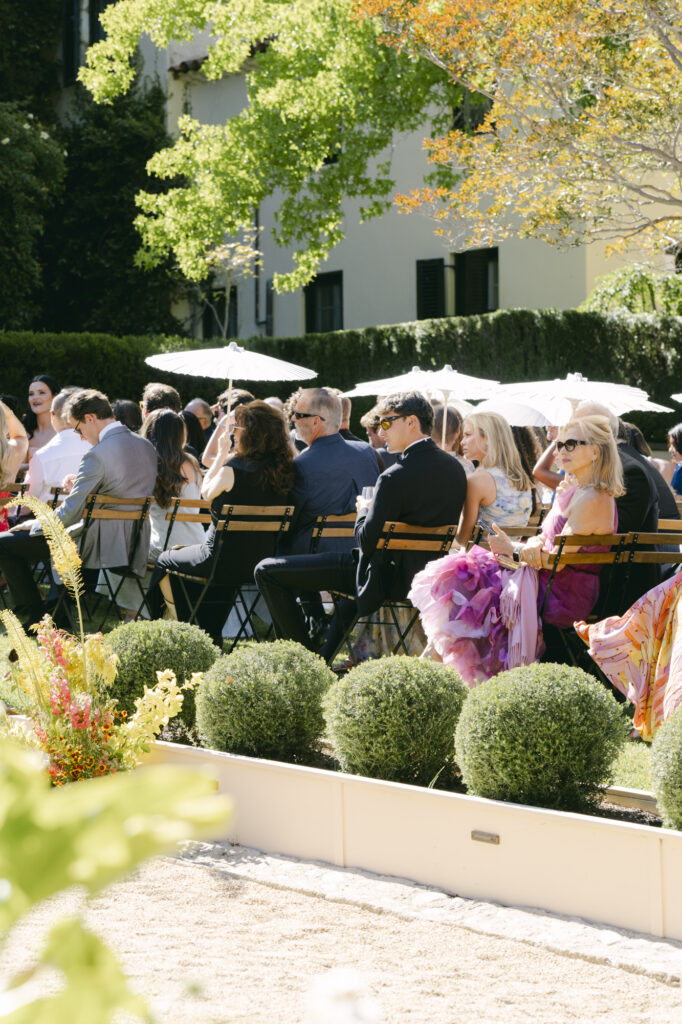 Wedding guests mingling and seated at bistro chairs under white parasols in the formal garden at Chateau Noel at Stonepine Estate, boxwood topiaries and the villa visible in the background