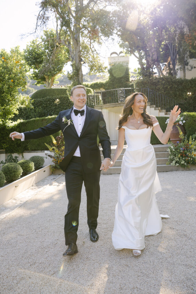 Bride and groom walking hand in hand and laughing through the grounds of Stonepine Estate in Carmel Valley, Chateau Noel visible in the background